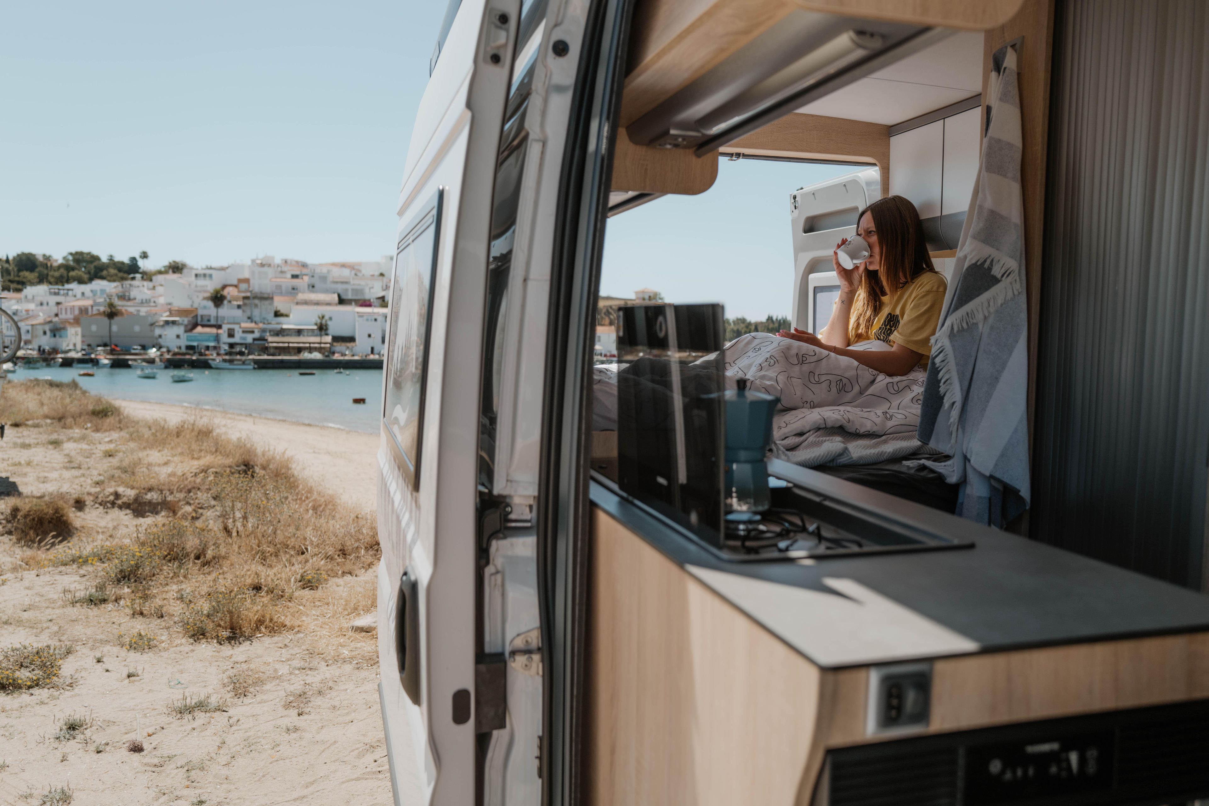 Elisabeth beve un caffè e dal letto del Campervan 540 Family guarda fuori la spiaggia e la città di Algarve in Portogallo