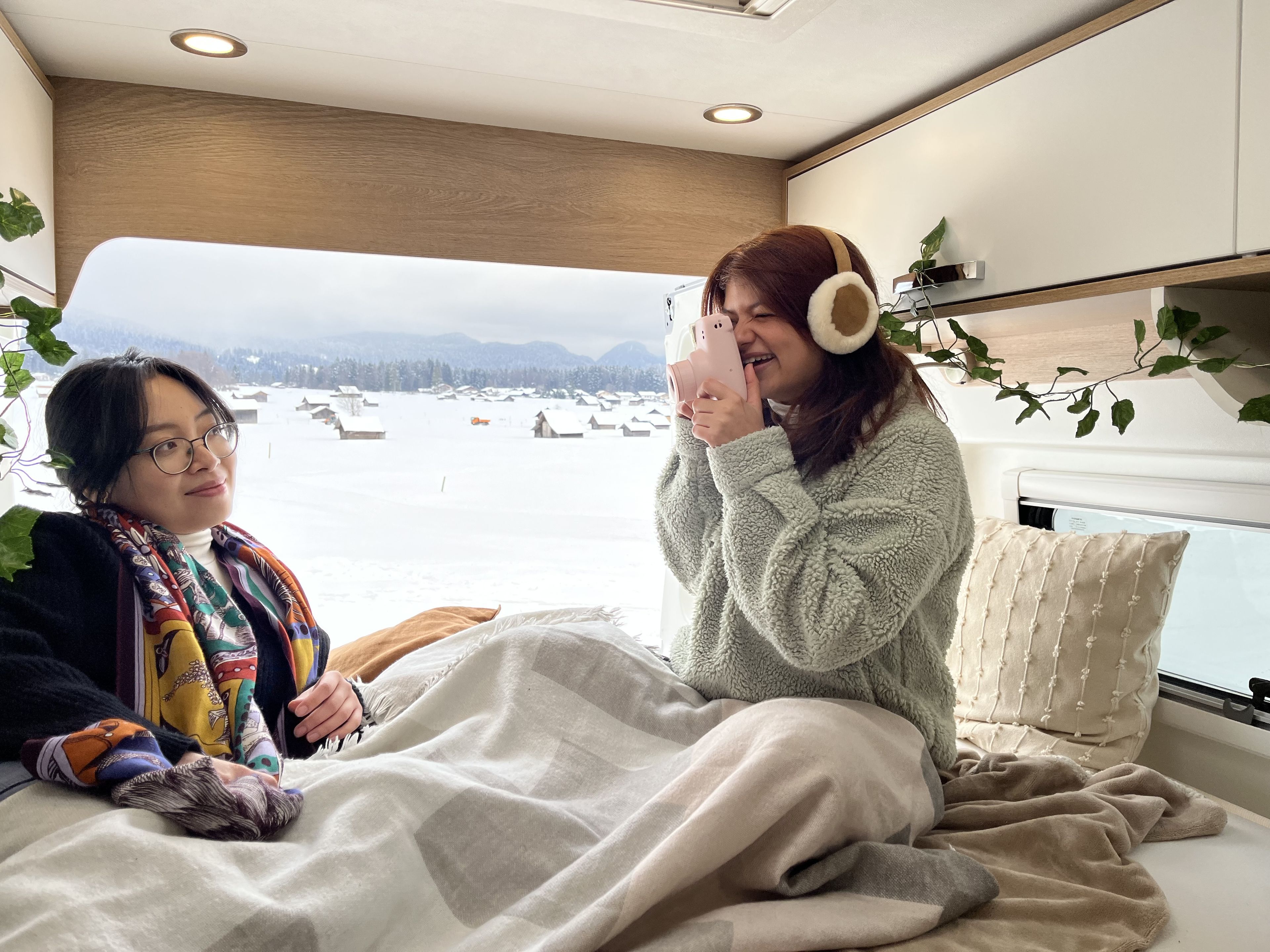 una ragazza che fotografa la sua amica con una macchina fotografica istantanea su un grande e comodo letto di un camper in un inverno innevato. Il letto è ben decorato con luci fiabesche, piante finte, coperte e cuscini caldi