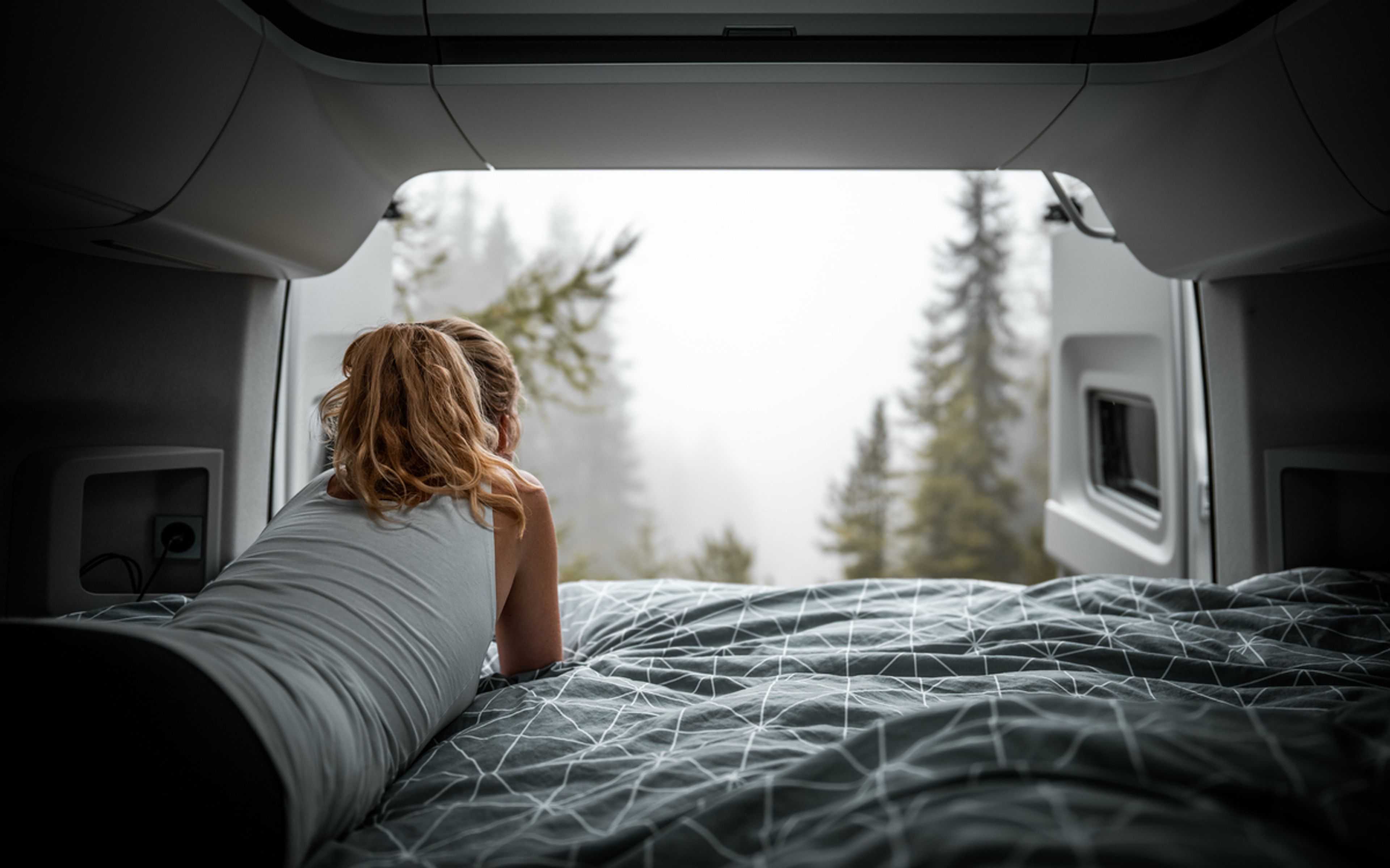 A woman laying comfortably inside the camper van enjoying the view of pine trees and foggy mountain