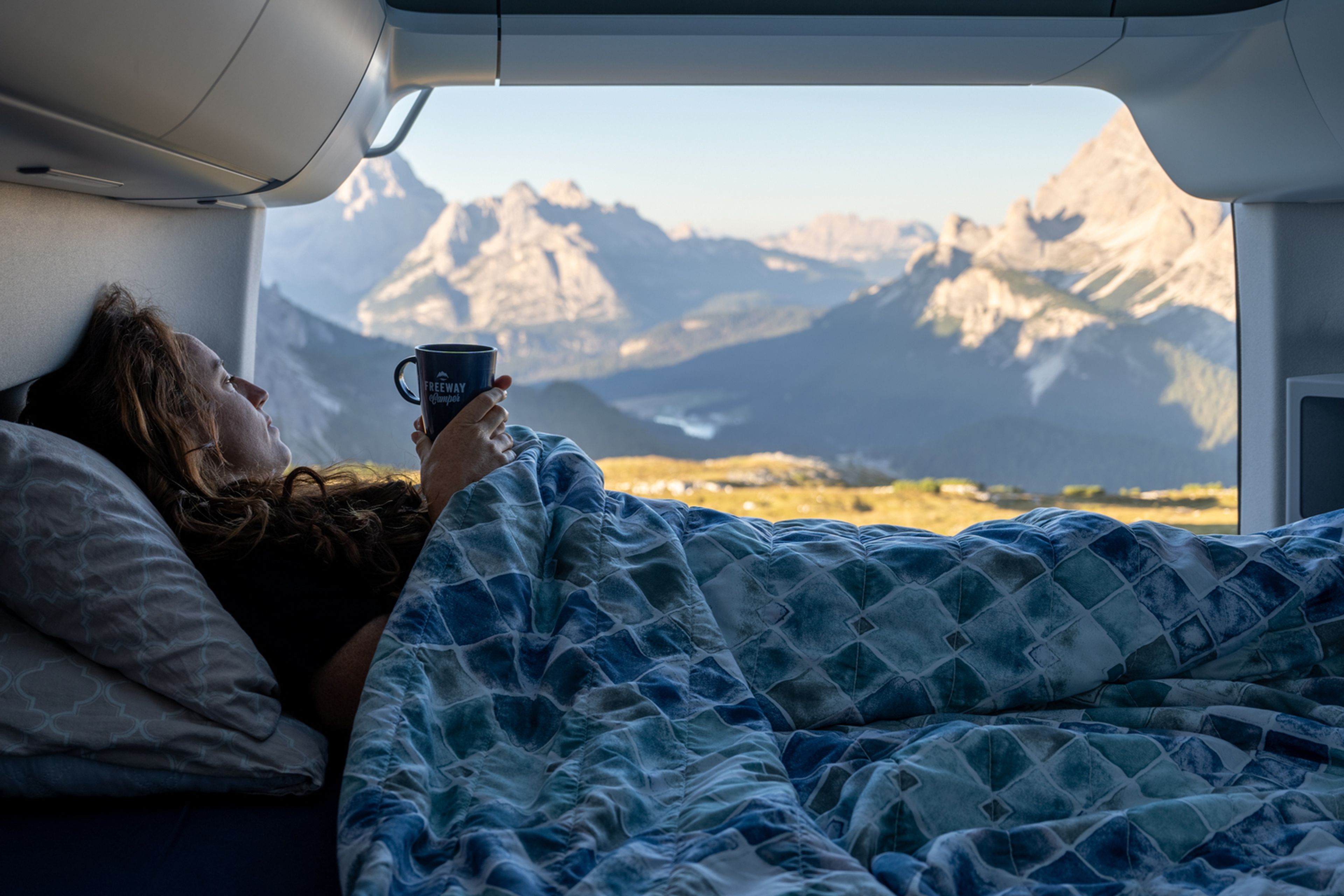 Una signora si sveglia con il caffè in mano. Si gode la vista mattutina delle Dolomiti sul letto di un camper.
