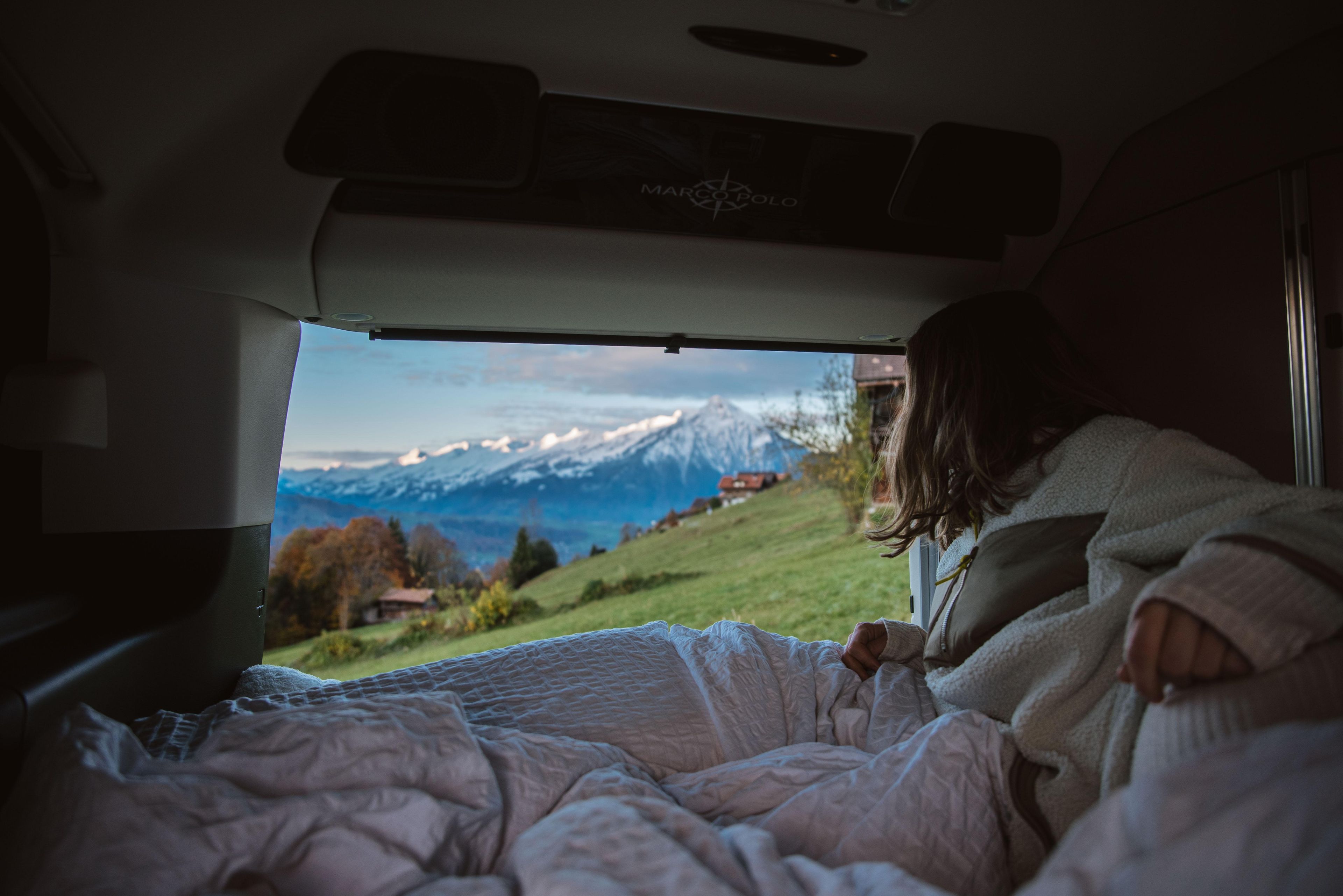 Una ragazza è sdraiata comodamente sul letto del VW Bulli Ocean T6.1 e guarda il paesaggio delle montagne e dei prati verdi della Svizzera
