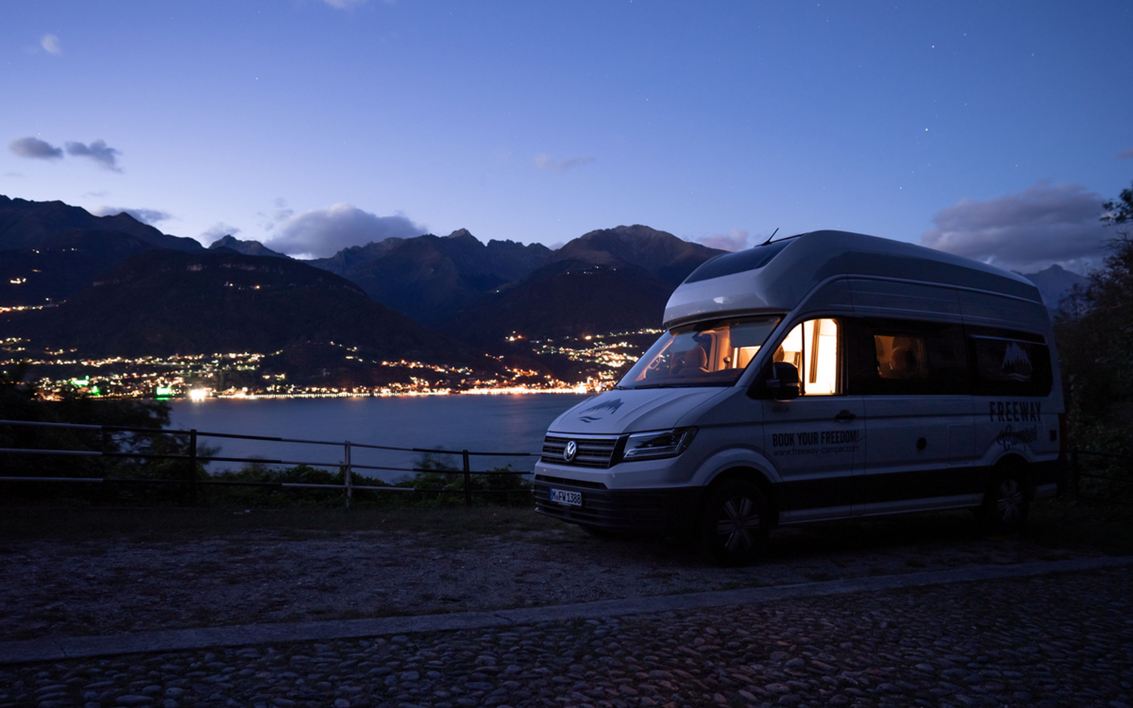 VW Grand California parks at night on the side of the road in Italy overlooking a city by the lake filled with lights