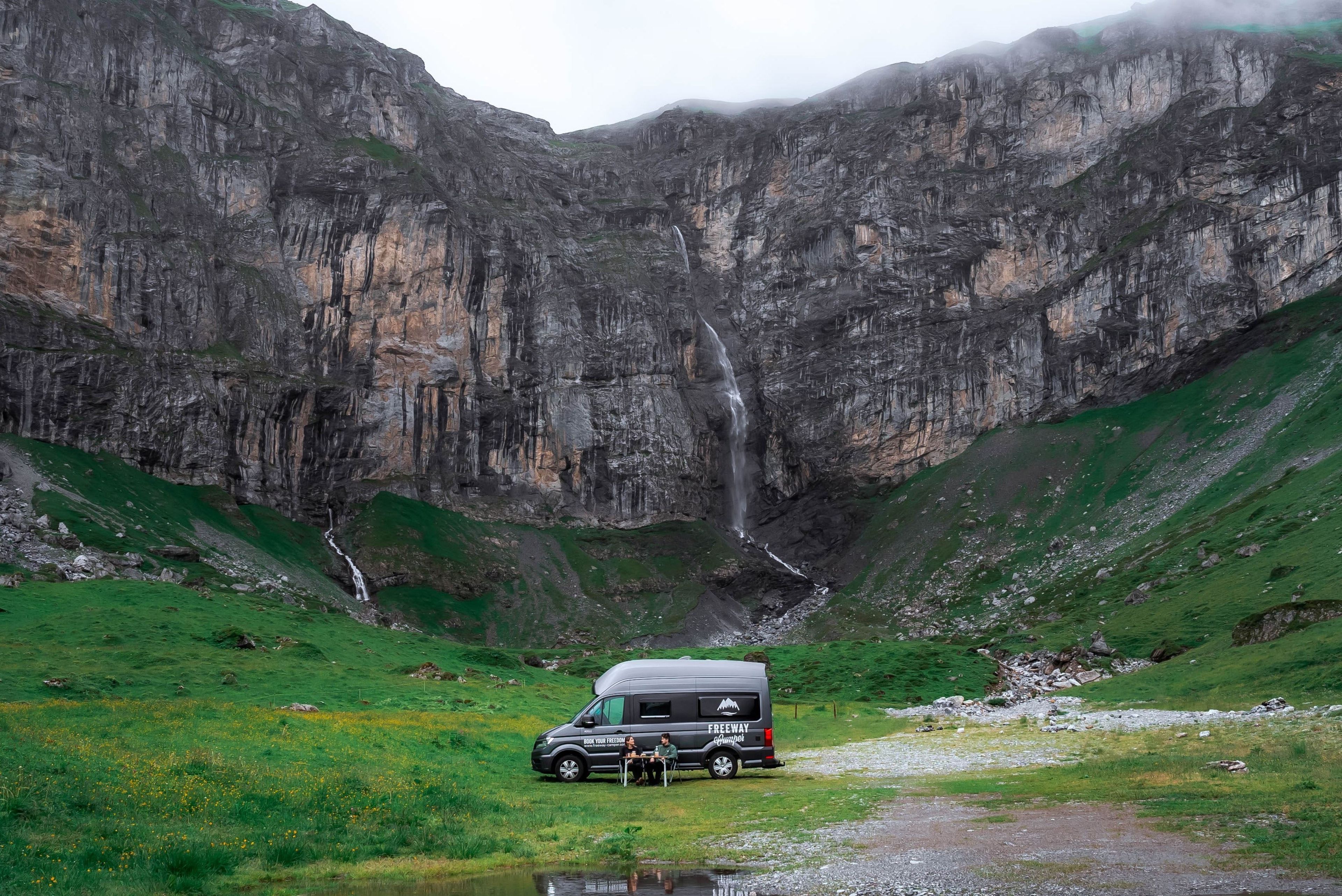 Vor einem Wasserfall in den Bergen sitzt ein Pärchen vor einem Campervan.
