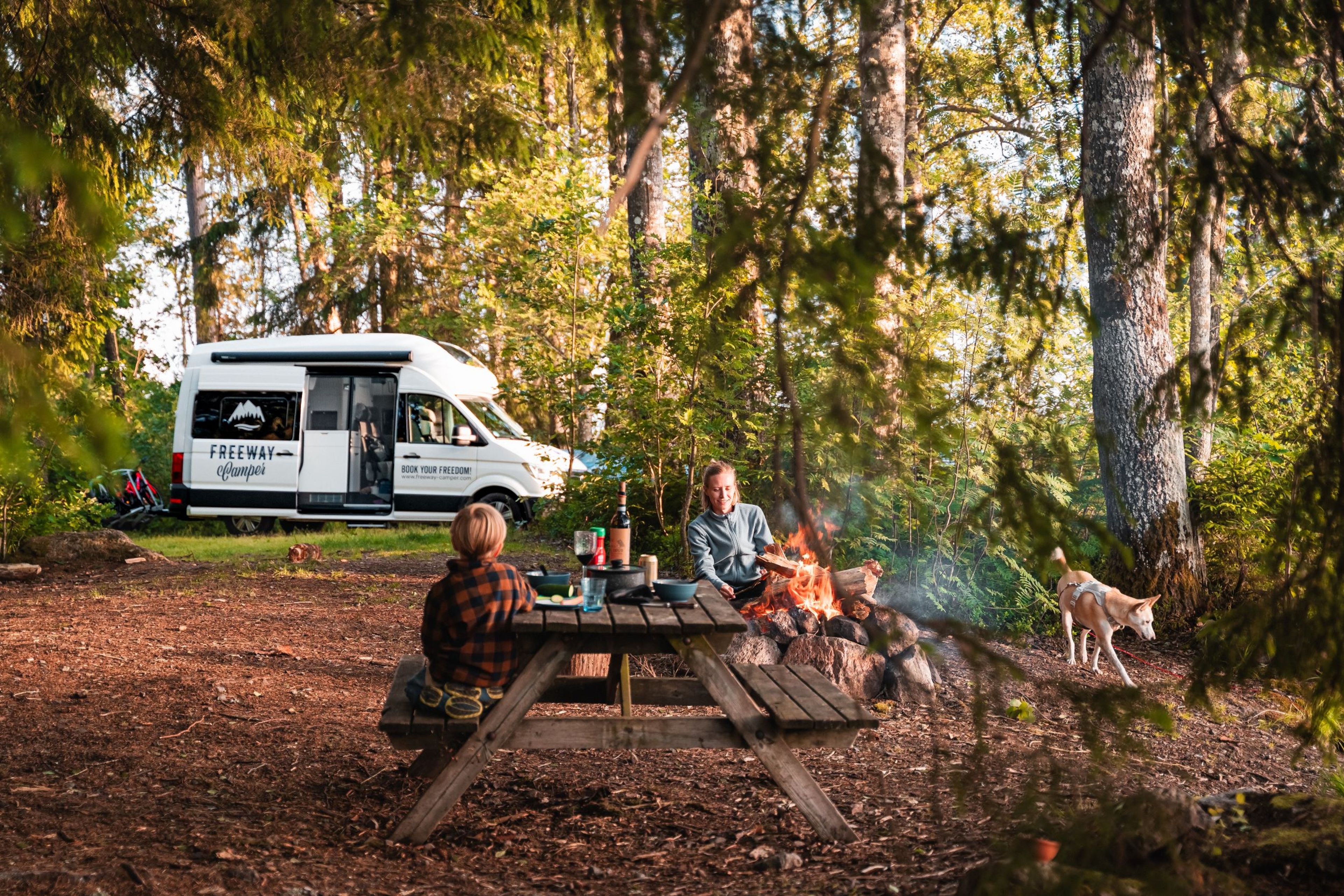 Familie sitzt zusammen bei Lagerfeuer auf Campingplatz im Wald