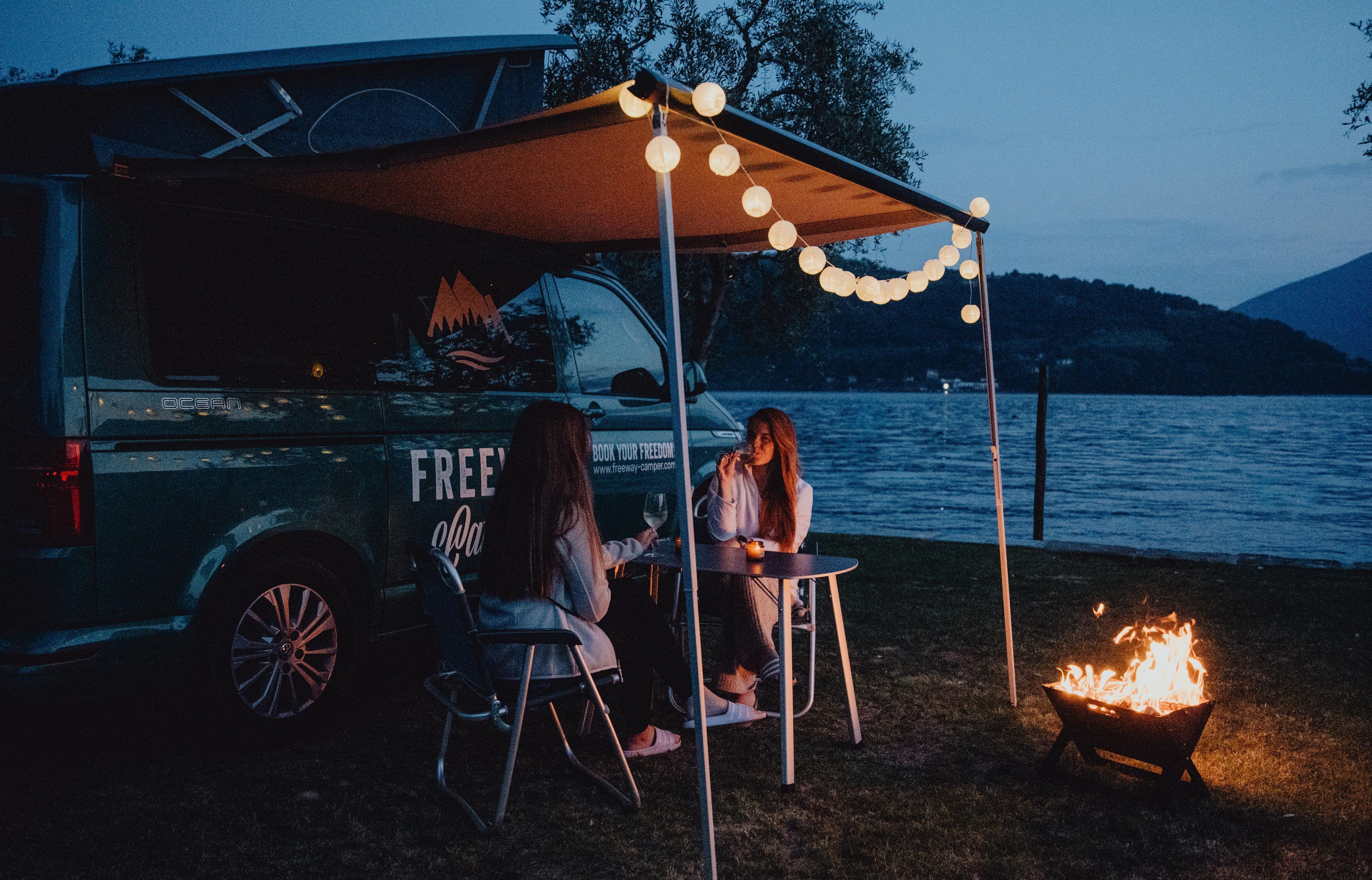 Due signore che si godono una serata accogliente accanto a un fuoco da campo con luci fiabesche appese al camper VW Bulli California Ocean T6.1 sul lago di Como, Italia