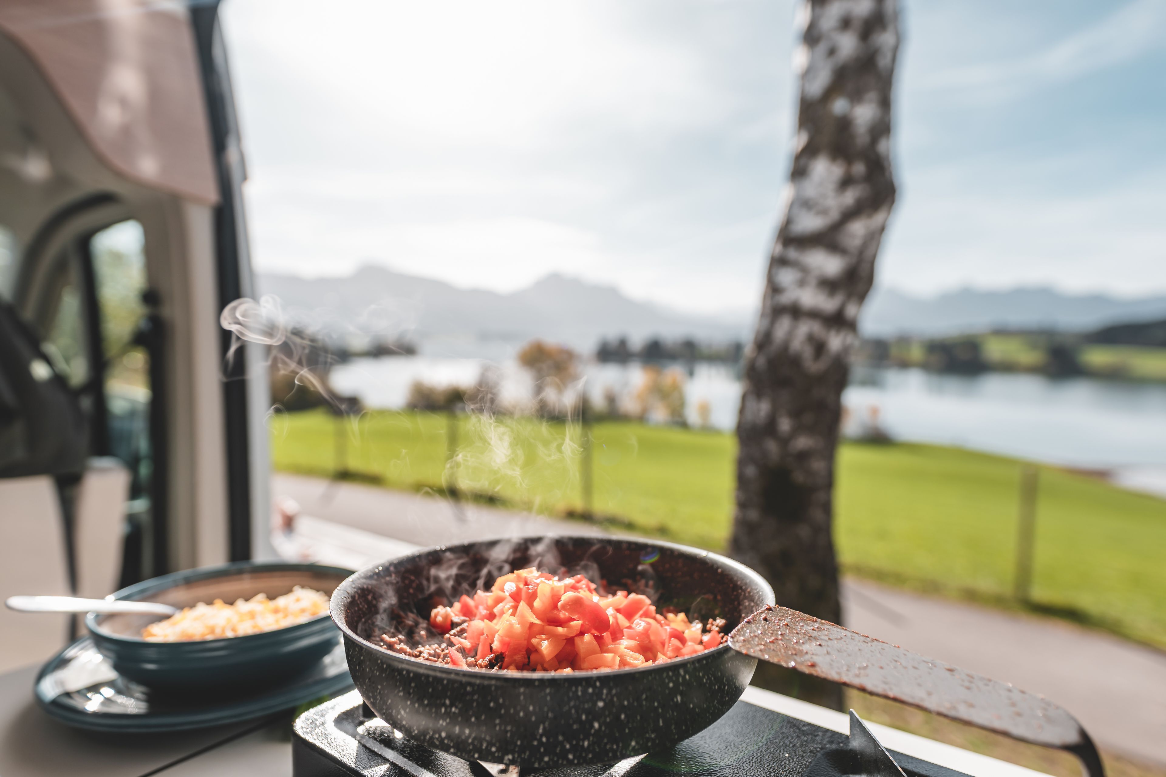 Essen wird auf einem Brenner im Wohnmobil gekocht, mit Blick auf den See und die Berge