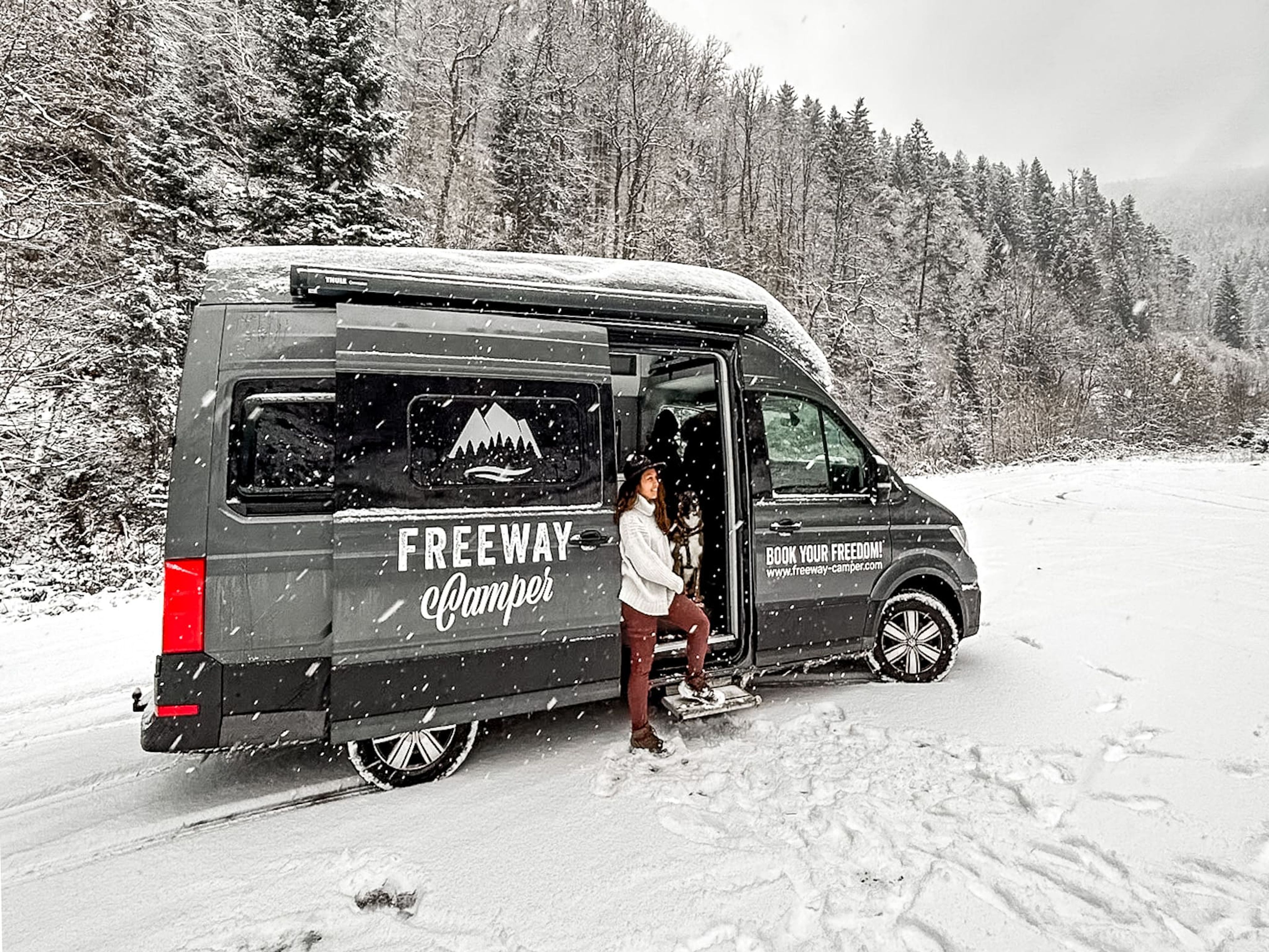 Viaggio in camper in Austria in inverno: una donna esce dal van sulla neve. Sullo sfondo gli abeti ricoperti da neve.