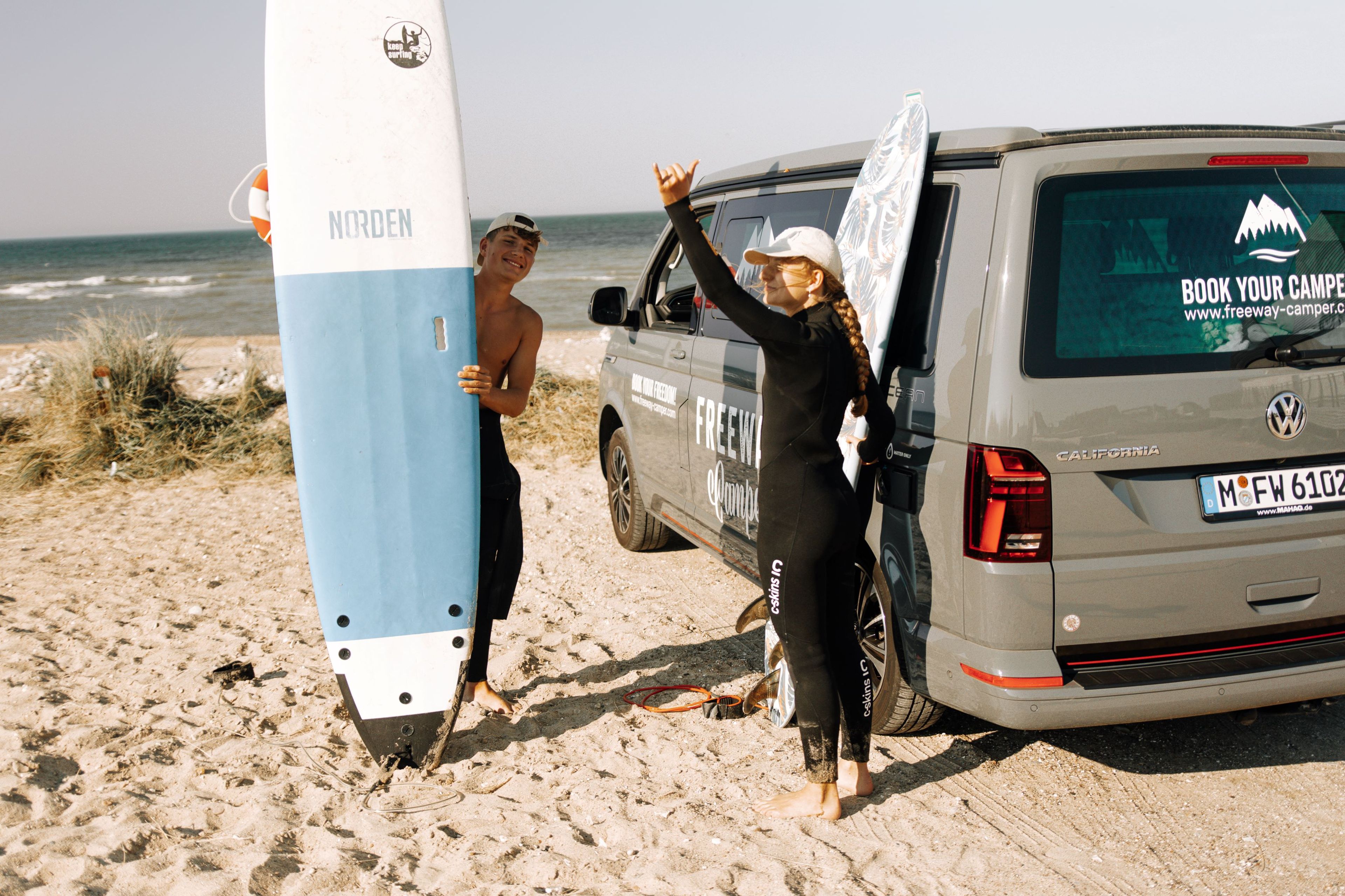 Zwei Personen haben Spaß mit VW Bulli und Surfboards am Strand in Dänemark