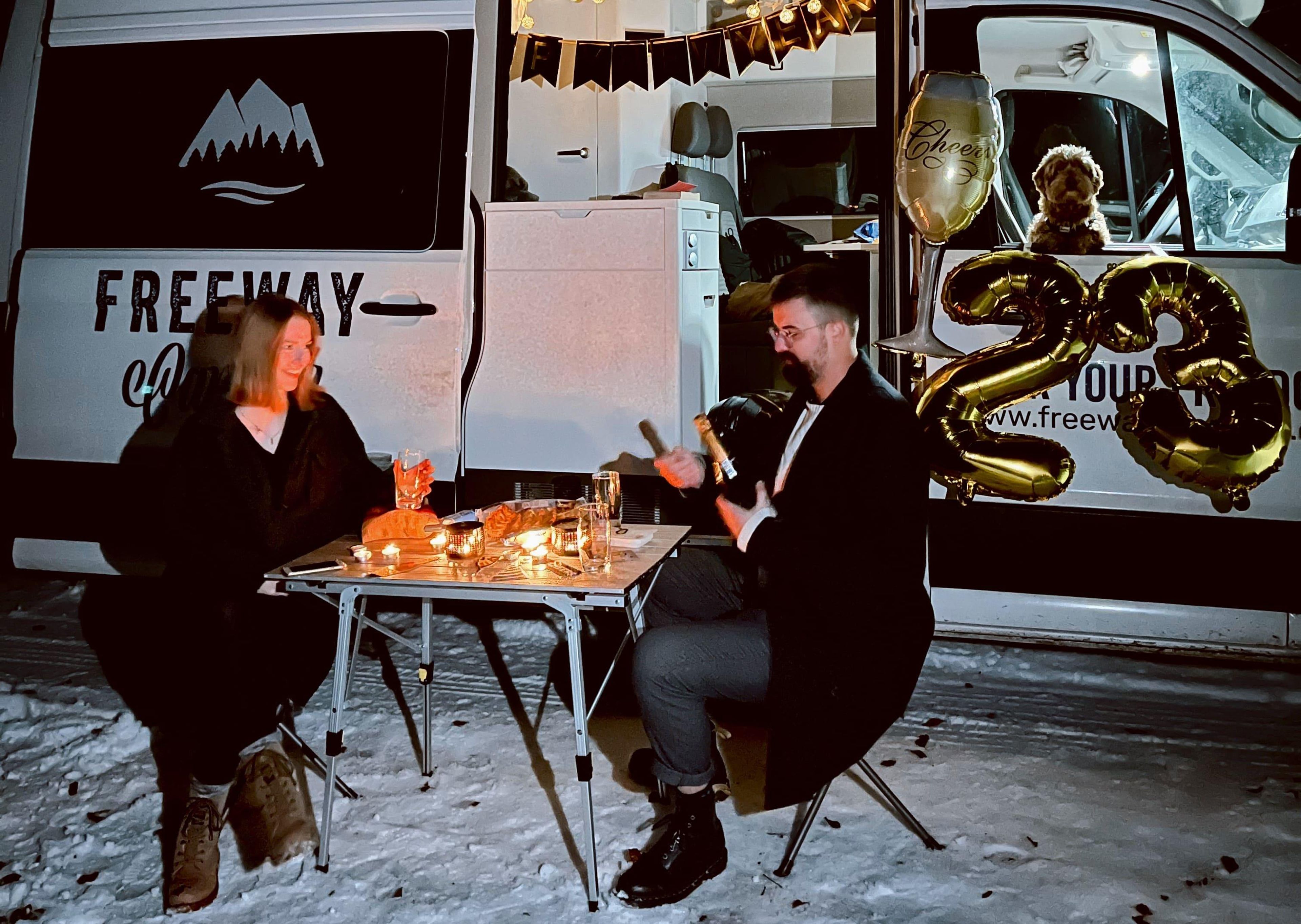 2 people sitting at the camping table in front of VW Grand California with New Year's Eve decorations