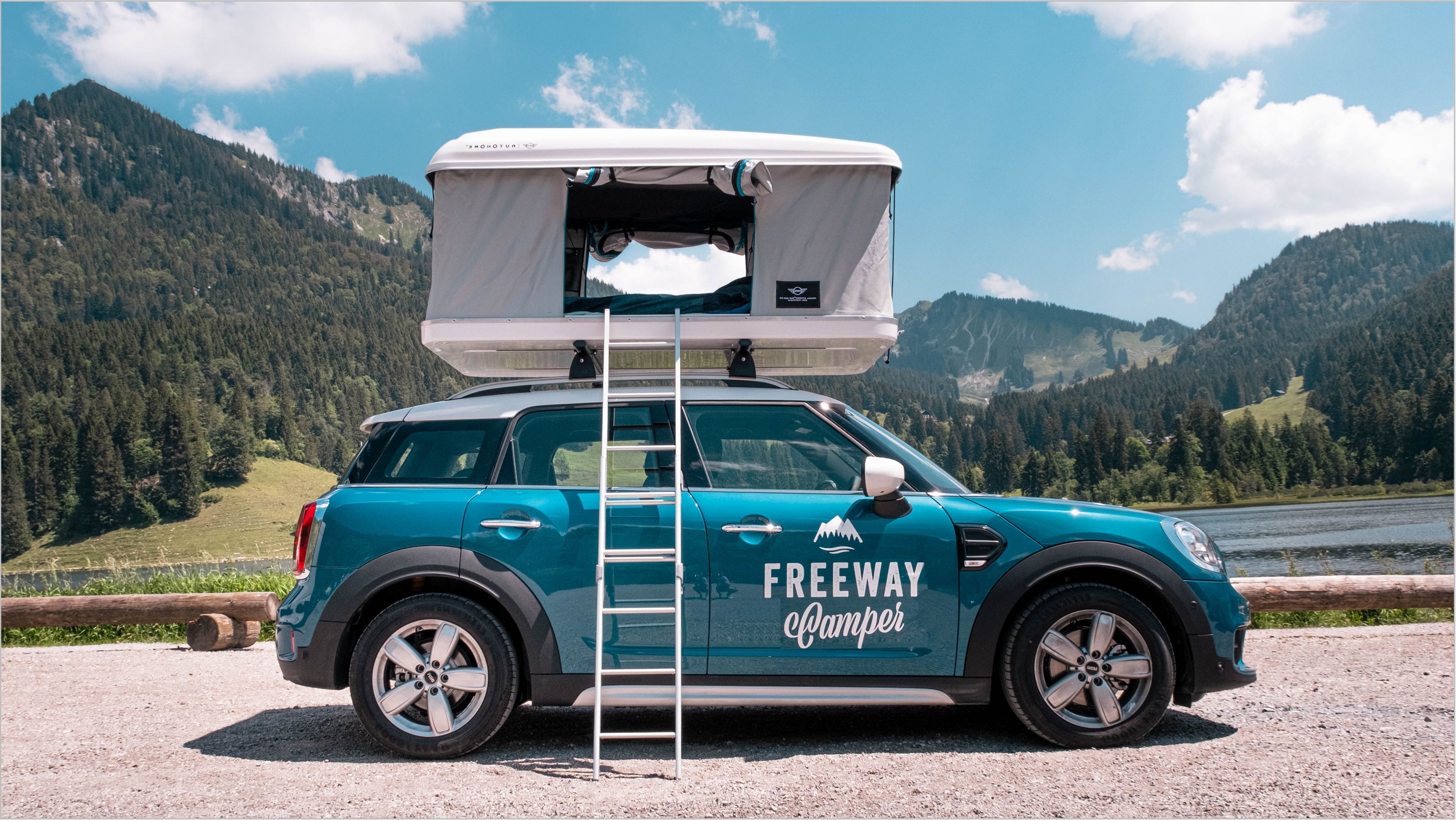 Blue FWC Mini Countryman with roof tent stands in the mountains by a lake.