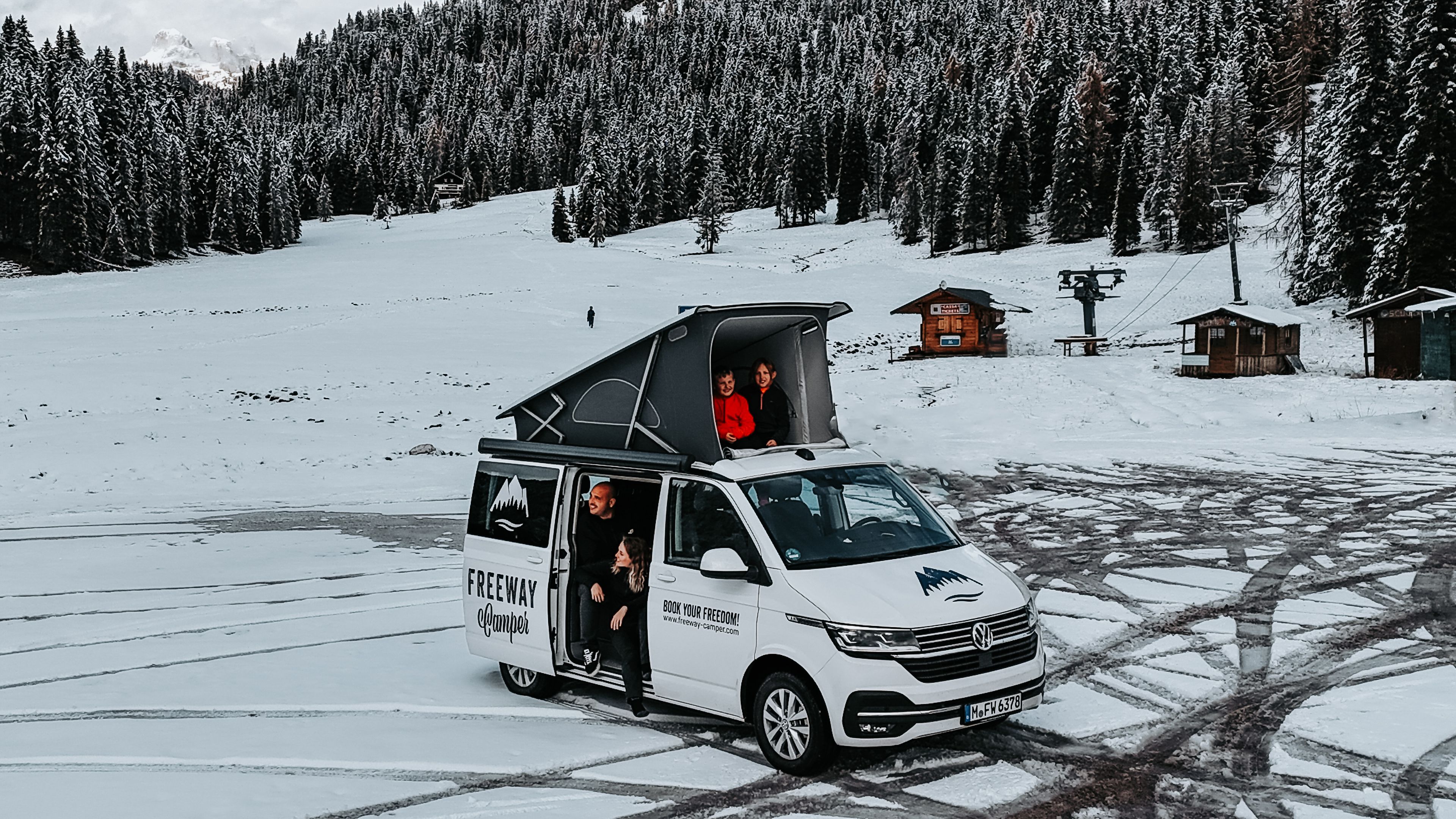 una famiglia di 2 bambini seduta all'interno del camper VW California Ocean con tetto apribile durante l'inverno nelle montagne del Nord Italia