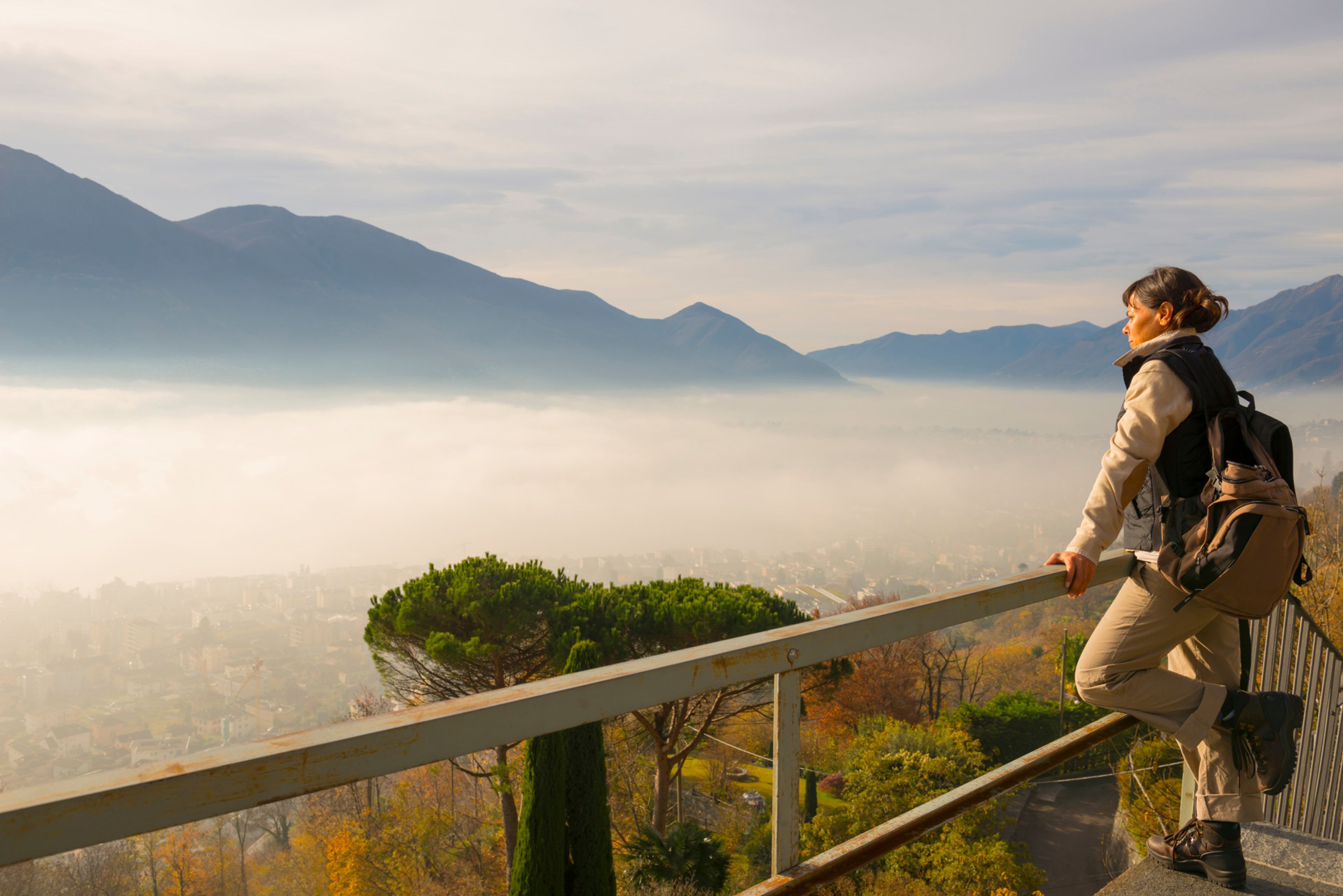 Donna con zaino in spalla che si appoggia a una ringhiera e gode della vista panoramica sul Lago Maggiore e sulle montagne del Ticino, in Svizzera.
